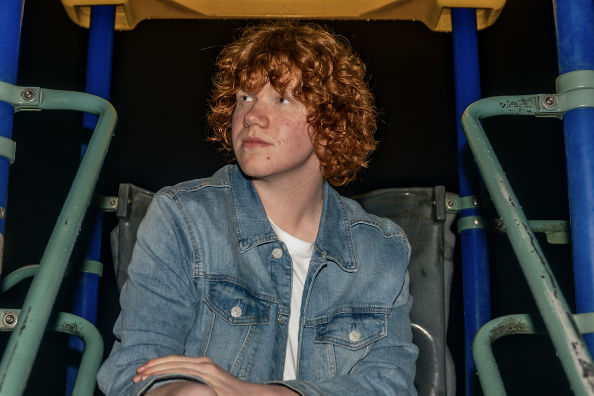 South Australian musician Ben Sutto with curly red hair wearing a denim jacket, sitting on playground equipment looking off-camera.